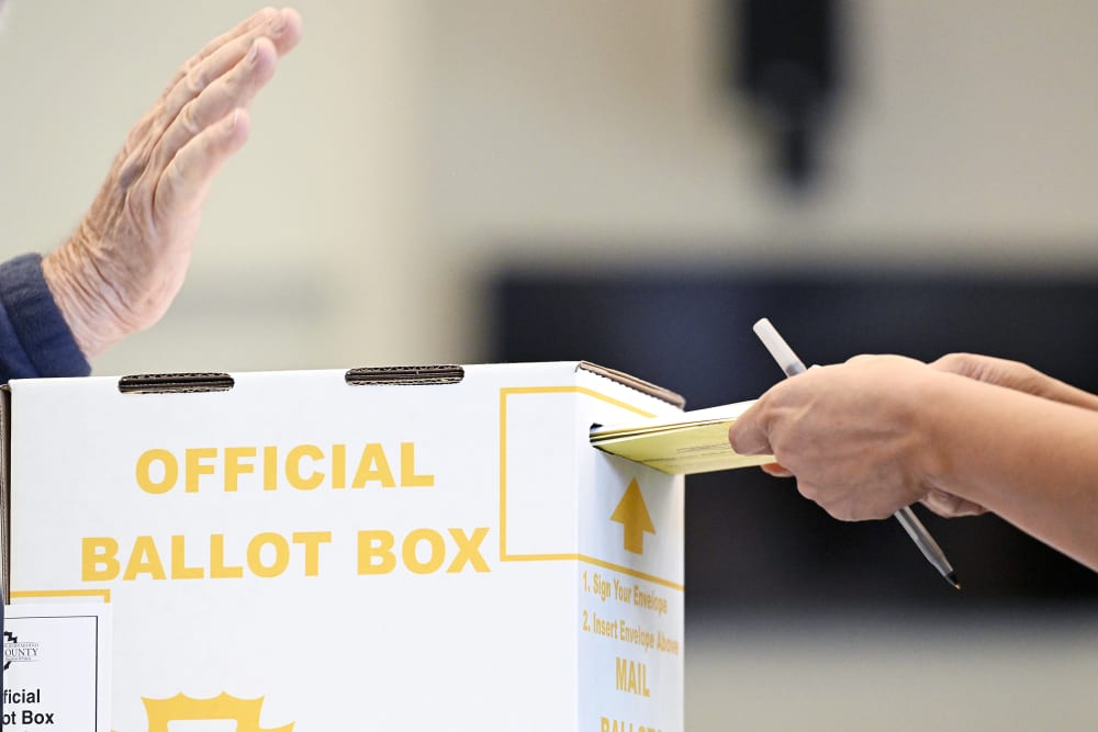 In a close-up shot, a person is putting their ballot in a box while an election official raises his hand.