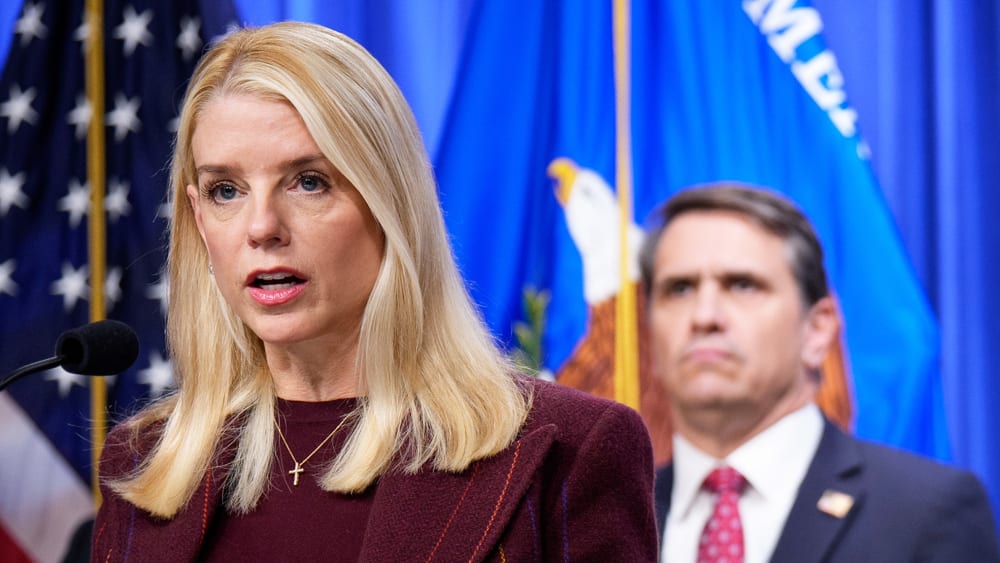 Pam Bondi stands behind a podium and microphone; Todd Blanche stands behind her, out of focus.