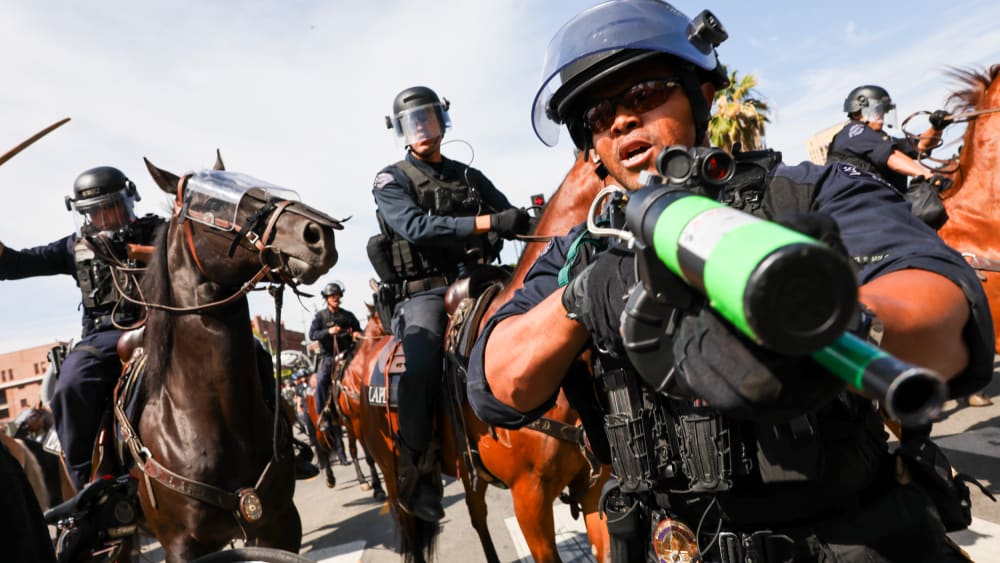 A police officer holds a weapon to the camera as protesters face off with police outside of a federal building in downtown Los Angeles for an anti-Trump "No Kings Day" demonstration.