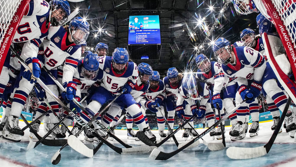 Hockey players in jerseys that read "USA" gather at the goal post. The photo is taken from a camera inside the goal.