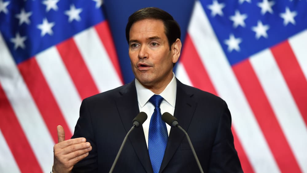 Marco Rubio is seen speaking in front of two US flags.