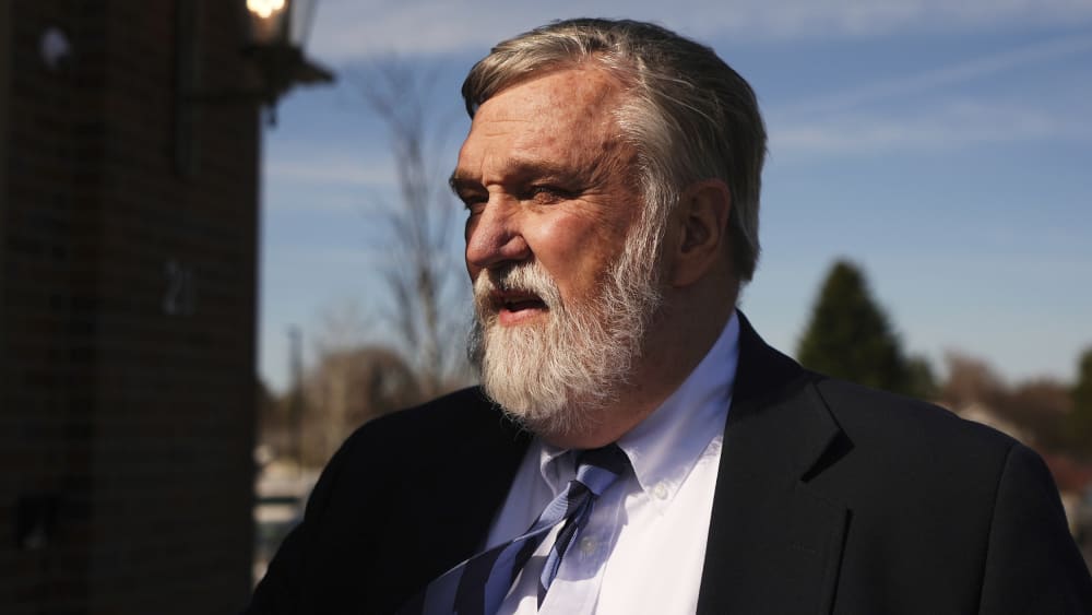 Doug Wilson stands next to a wall facing the sun, his eyes are squinted.