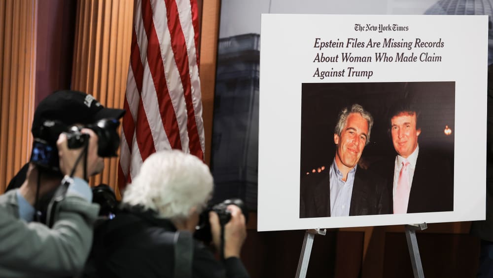 There are two photographers on the left bottom of the frame taking pictures of a poster that shows a photo of Donlad Trump and Jeffrey Epstein together.