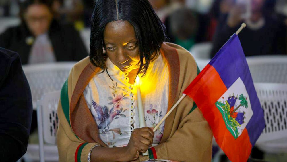 Haitian community activist bows her head down as she prays and holds a candle and Haiti flag.
