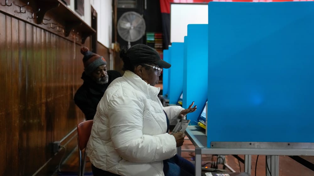 A female voter sits in front of a voting machine while a man can be seen in the background.