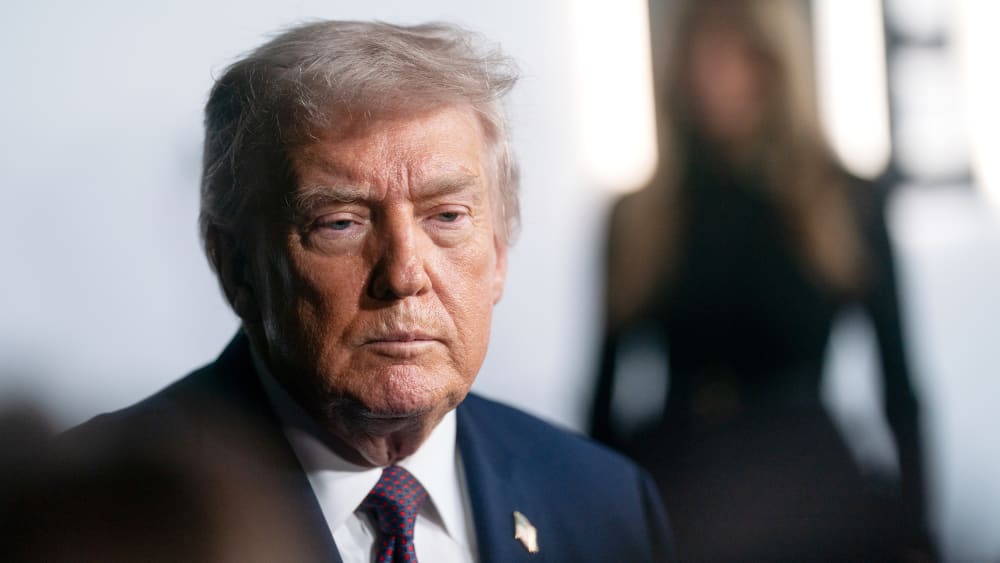 A tight shot shows Donald Trump looking at the reporters and Melania Trump can be seen in the background very out of focus.