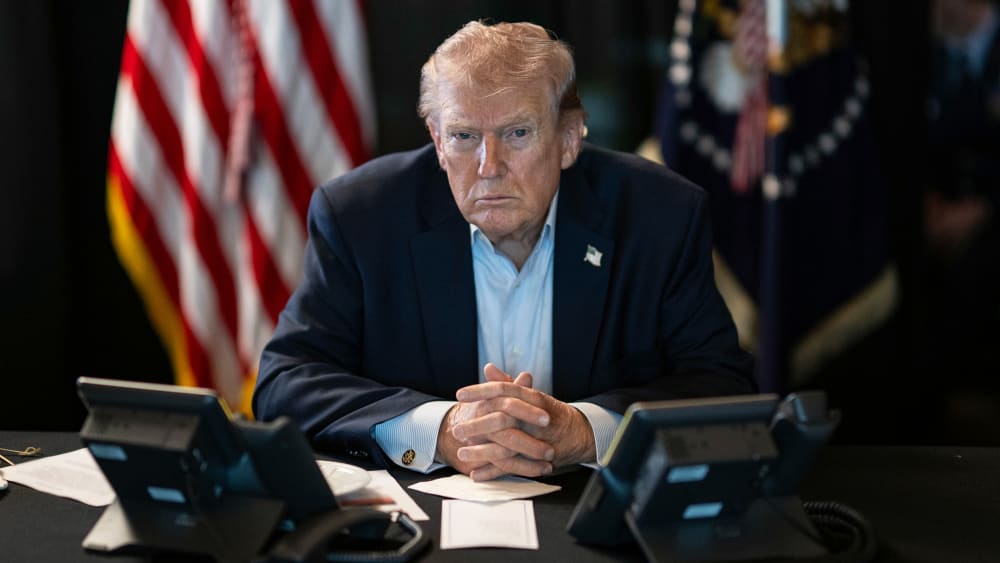 Donald Trump sits at a table with two phones in front of him and U.S. flags behind him.