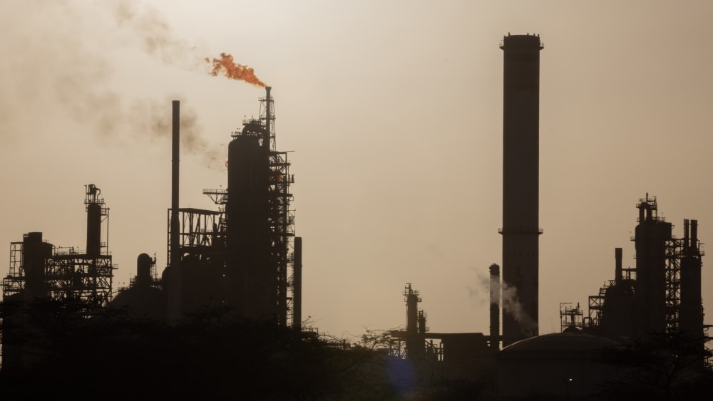 An oil refinery in Venezuela is seen in the distance while smoke and fire is coming out on the left side of the frame.