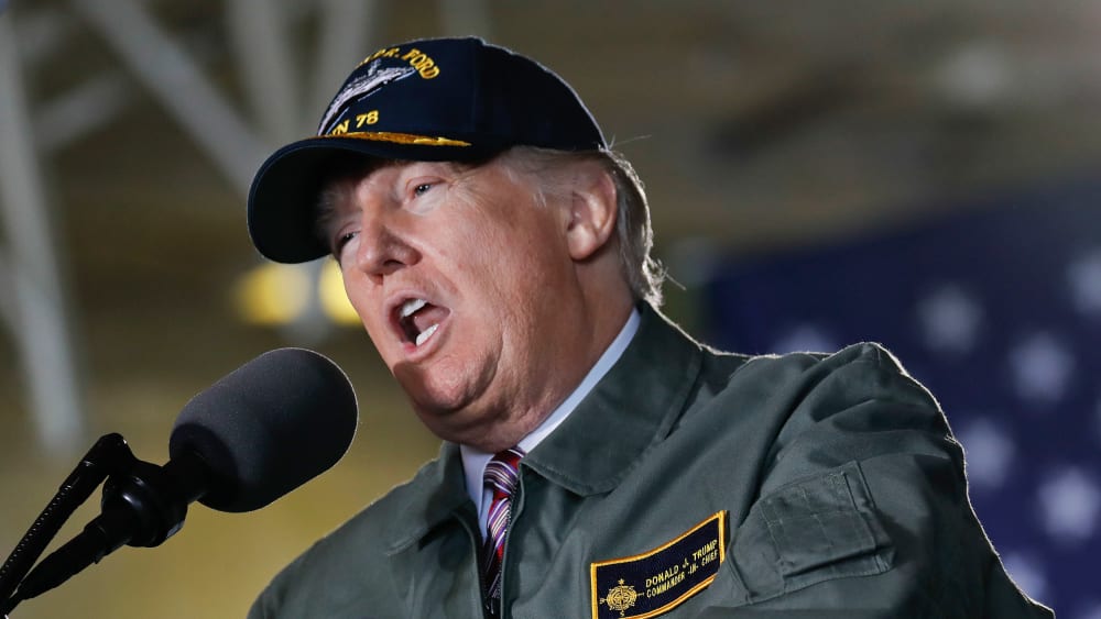 Donald Trump speaks into a microphone while wearing a USS Gerald R. Ford jacket and hat.