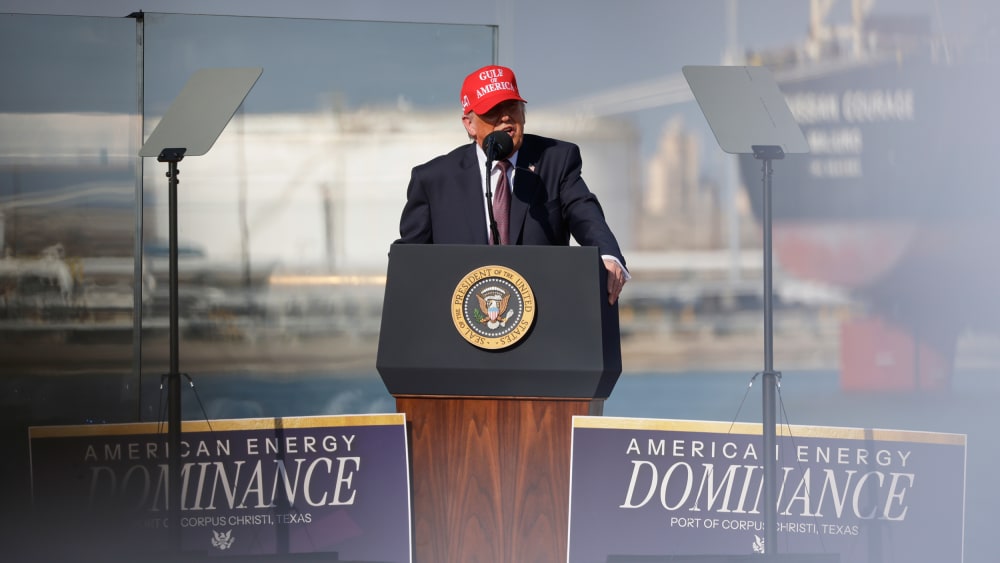 Donald Trump speaks into a microphone at a podium. He's wearing a red hat that says "Gulf of America."