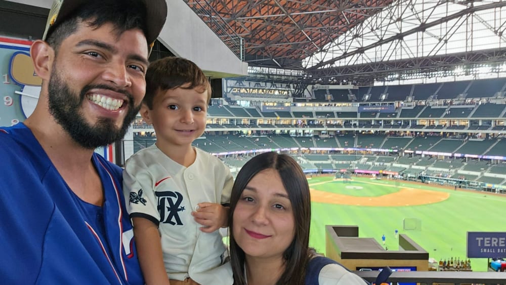 A man, woman, and small child, all in baseball jerseys. Behind them is a baseball field.