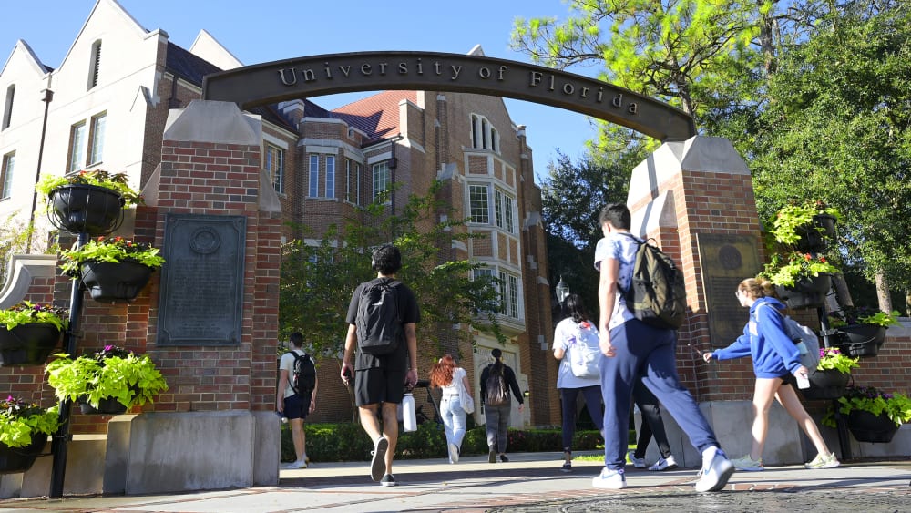 Students walk under the arch of University of Florida entrance on a sunny day.