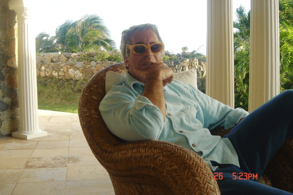 Jeffrey Epstein sits at a patio wearing yellow framed sunglasses.