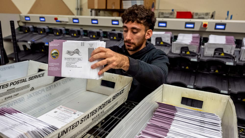 A person grabs a stack of mail-in ballots from a United States Postal Service tray of ballots.