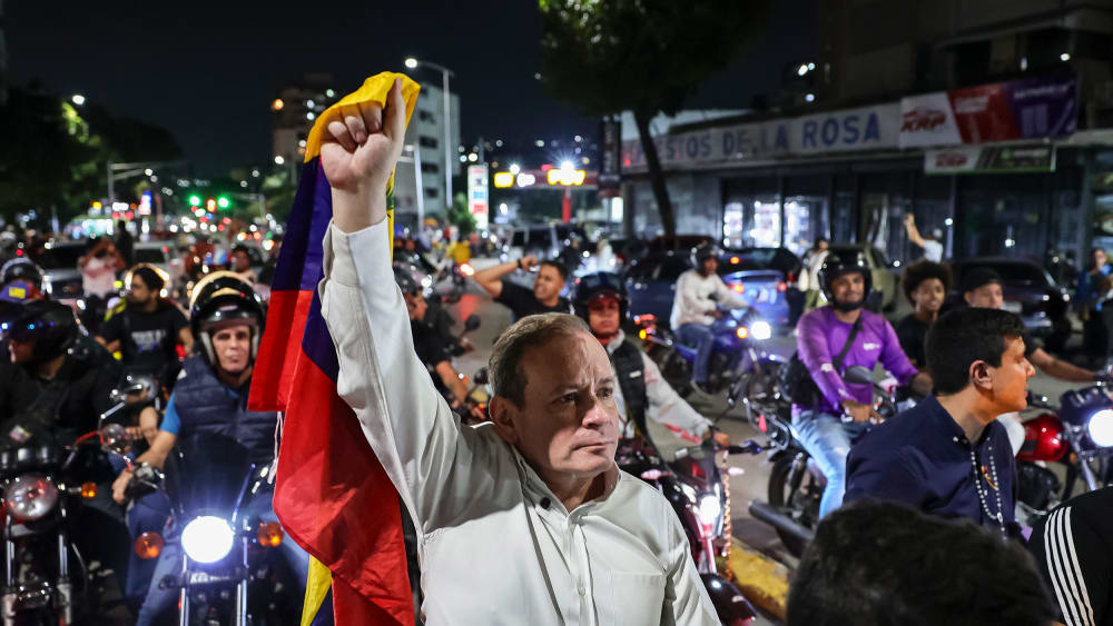 Juan Pablo Guanipa, center, stands in the street, holding a Venezuelan flag up with one hand. He is surrounded by people on motorcycles.