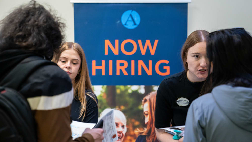 Two people talking with two job seekers in front of a &ldquo;Now Hiring&rdquo; sign.