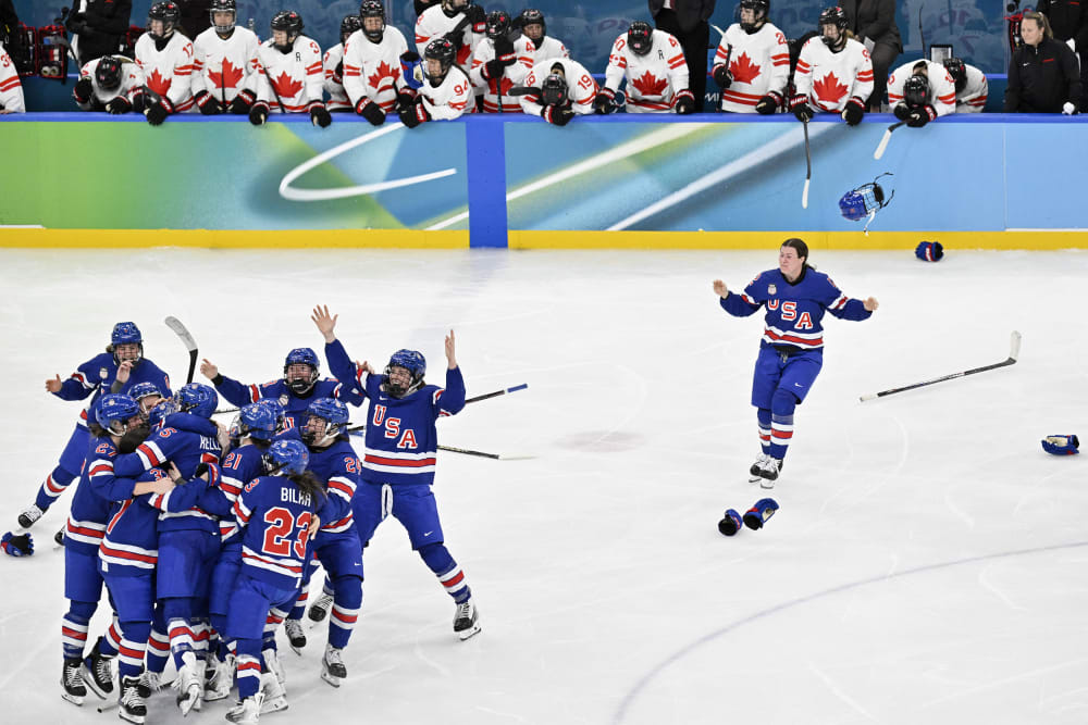 A group of US hockey players are huddled on the left side of the frame as one player runs toward them in celebration.