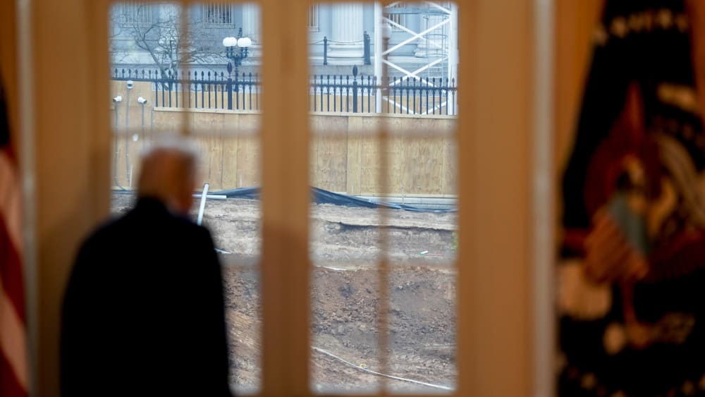 Donald Trump, out of focus in the foreground, looks away from the camera and out a set of windows, where construction is seen in focus.