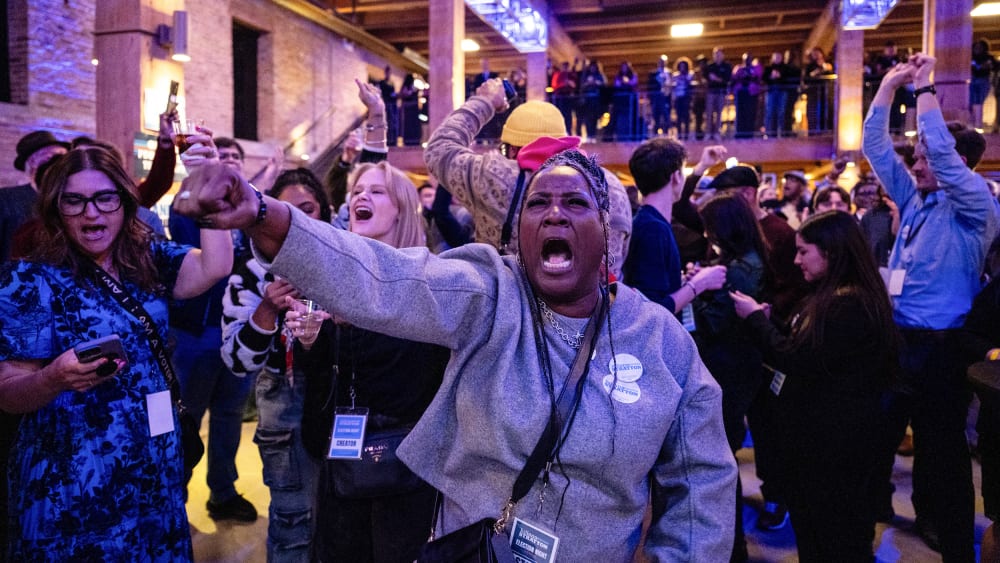 A woman screams with her fist up in a celebratory matter while other people doing similar stuff in the background.