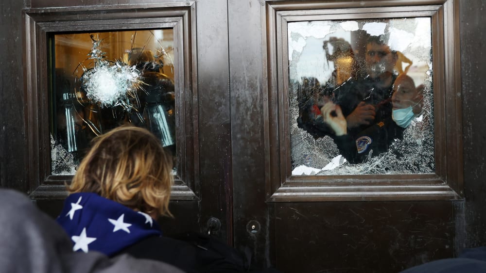 Police officer looks through a broken glass window as protesters stand outside.