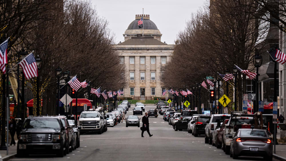 A street lined with cars leads up to the North Carolina State Capitol. A person crosses in the middle of the street, and American flags are on each of the lampposts.