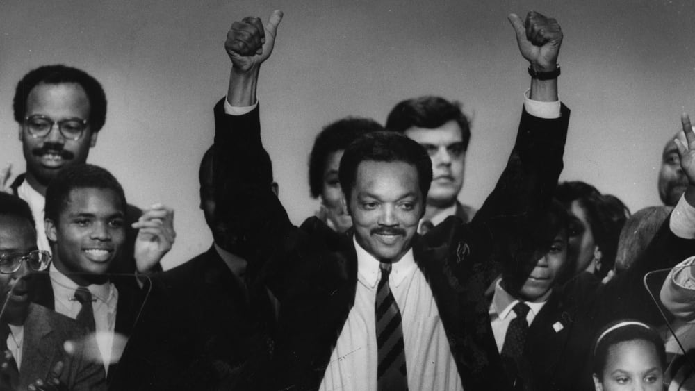 Jesse Jackson Sr. lifts his arms surrounded by a group of people mixed-aged people.
