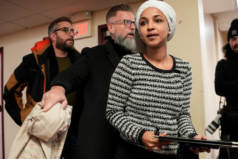 Ilhan Omar walks into a room for a town hall meeting, followed by two men who seem to be security personnel.