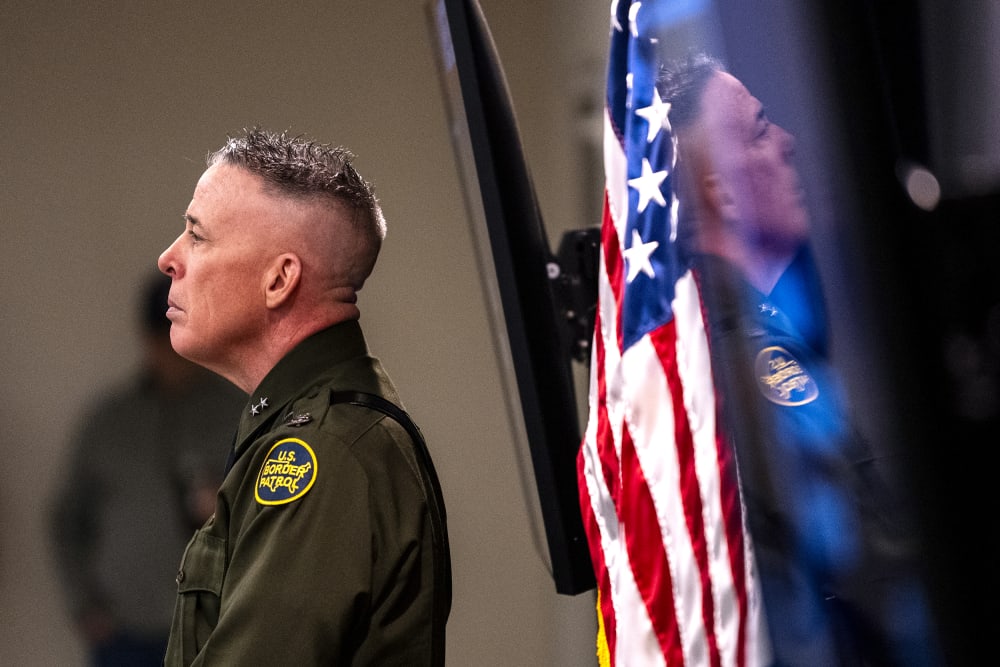 Greg Bovino is seen from the side while he looks on during a presser. His reflection can be seen behind him.