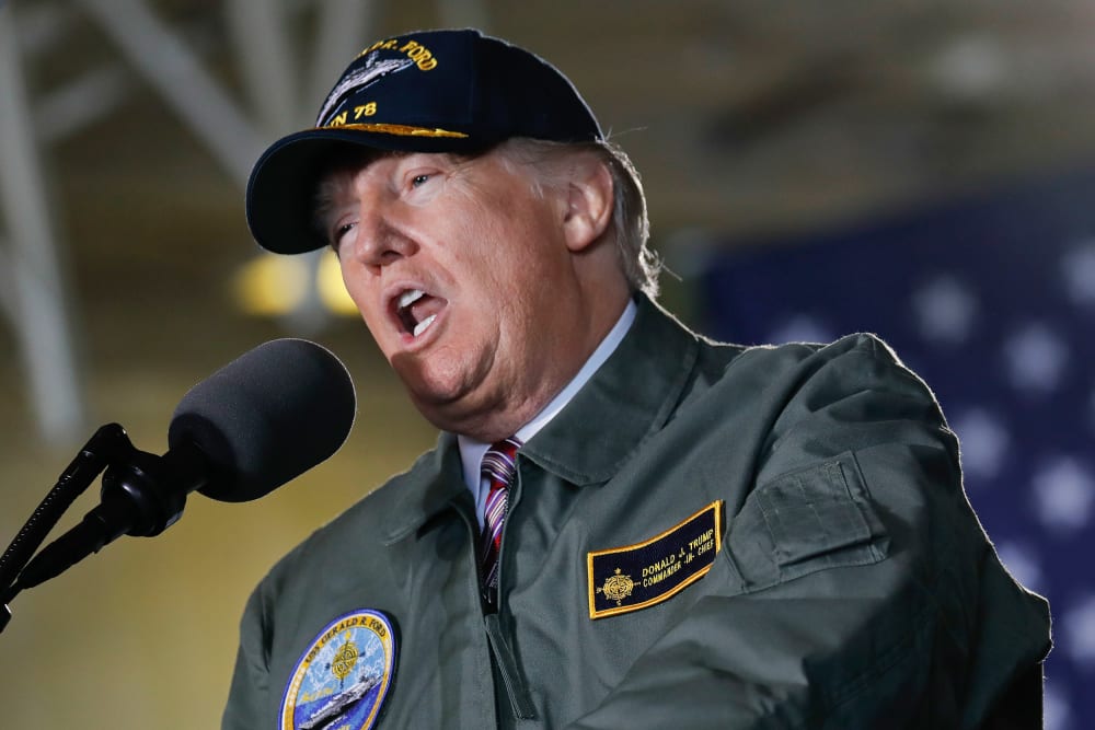 Donald Trump speaks into a microphone while wearing a USS Gerald R. Ford jacket and hat.
