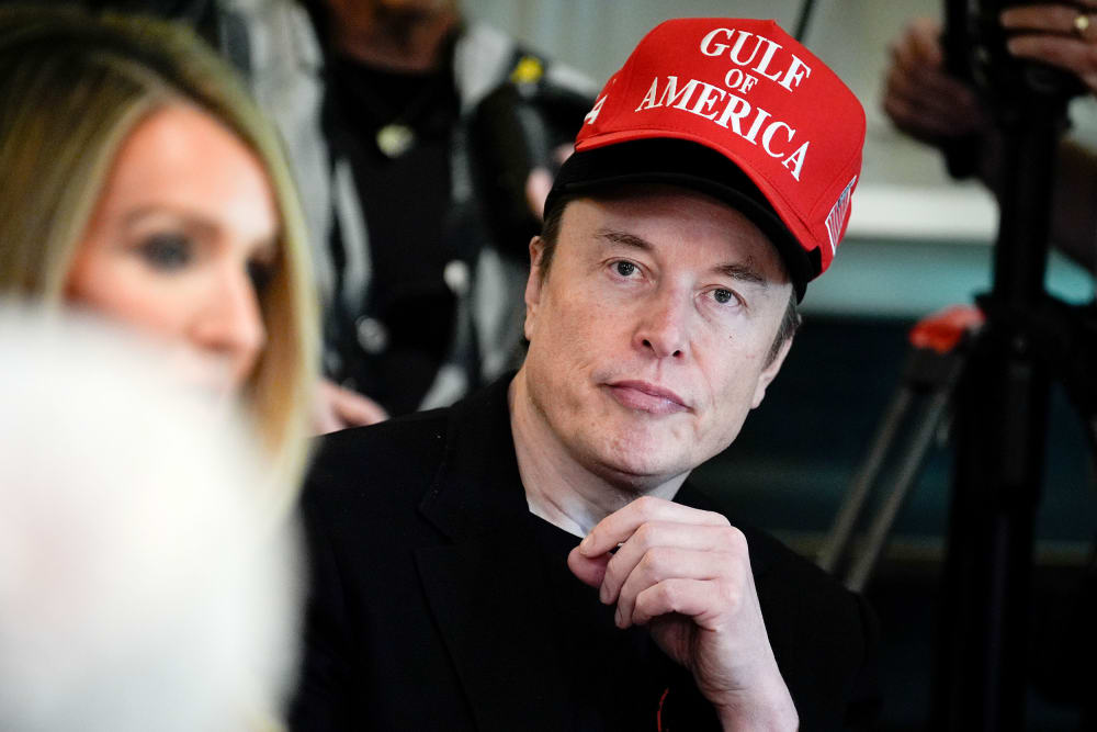 Elon Musk looks on while wearing a red Gulf of America cap over his black cap.