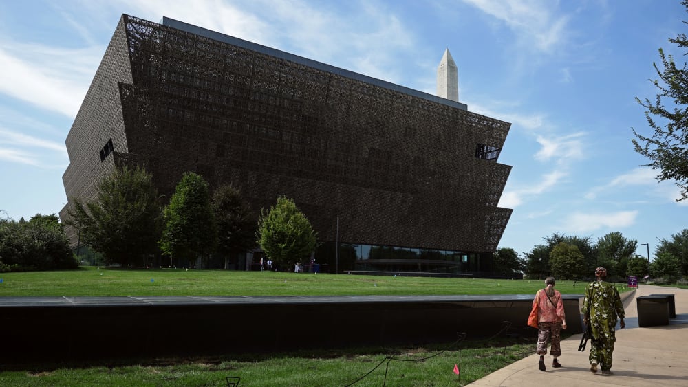 Two people walk by the National Museum of African American History and Culture on a sunny day.