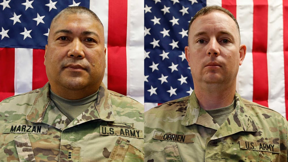 Two people in army uniforms in front of American flags.