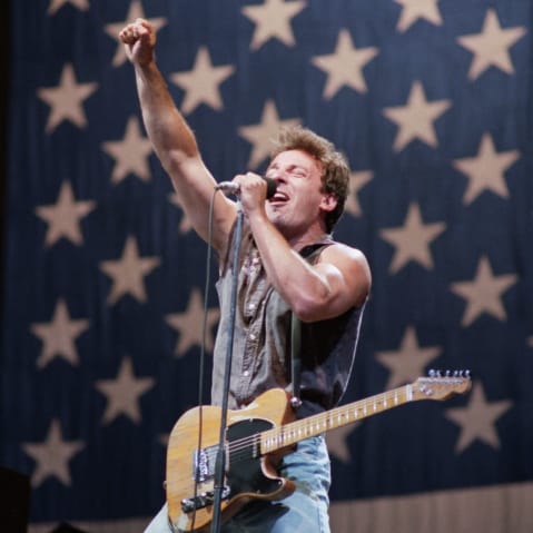 Bruce Springsteen with his guitar on stage, in front of a giant American flag.