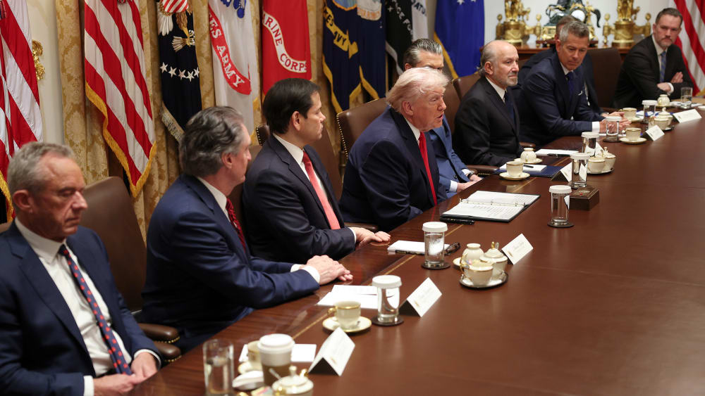 Donald Trump, sitting at the center of the group, speaks while his cabinet members to his left and right listen.