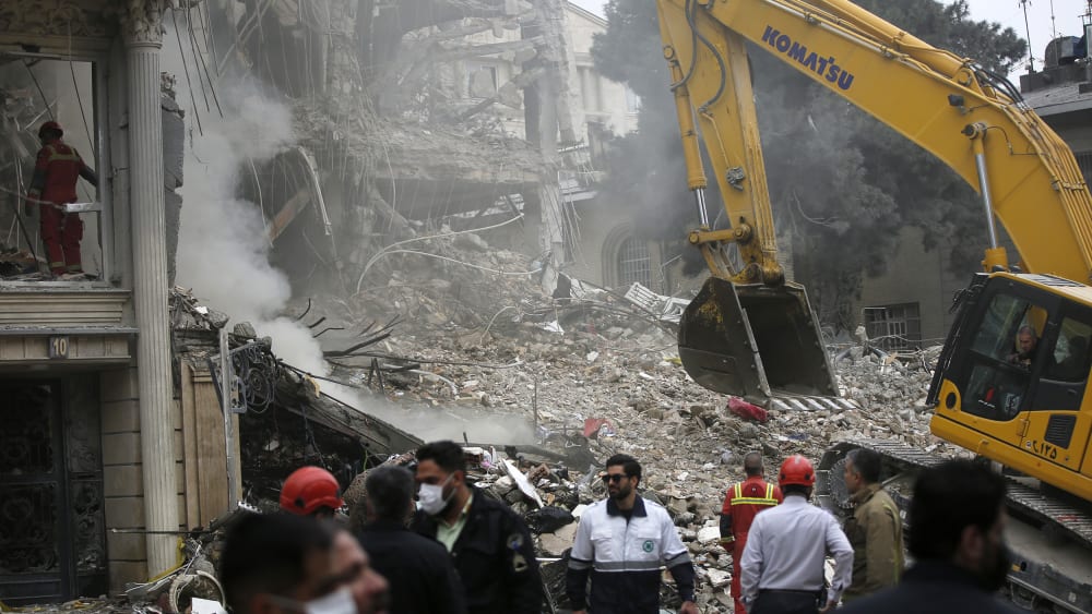 People are standing in front of a destroyed building as a work machine is seen in background.