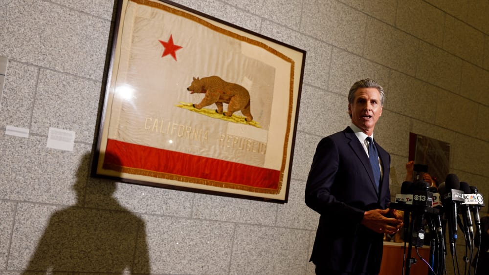 Gavin Newsom stands behind news microphones and in front of a framed California state flag.