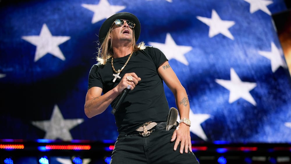 Kid Rock holds a microphone on stage. The background is an American flag.