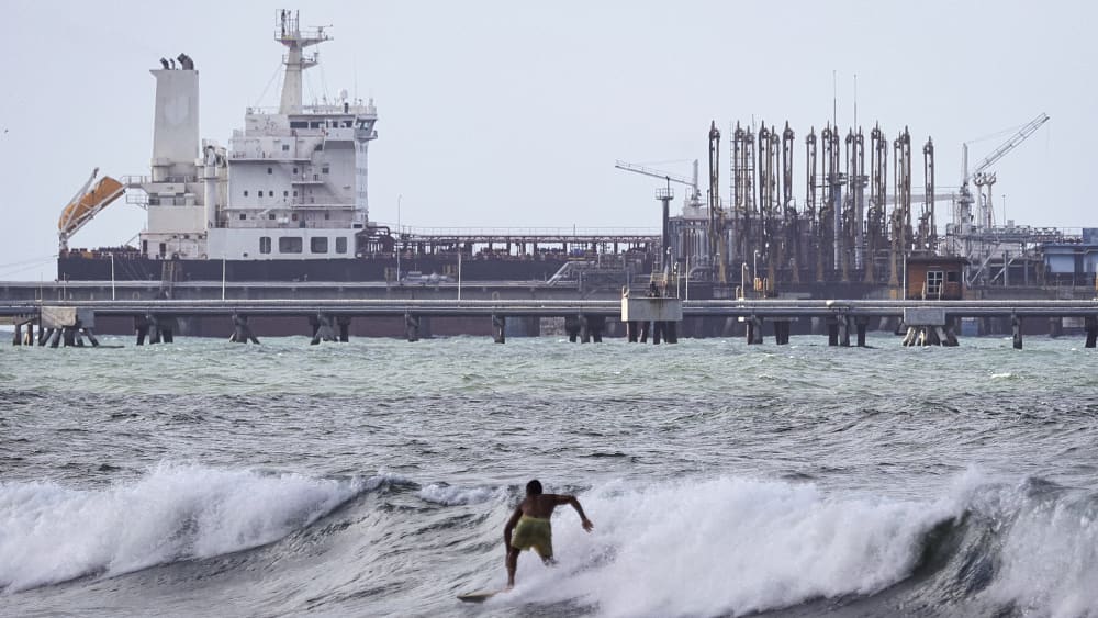 A man is seen from behind as he surfs in front of an oil tanker in the distance.