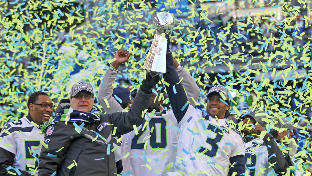 Paul Allen, left, and Russell Wilson hoist the Lombardi trophy in the air as blue and green confetti falls around them.
