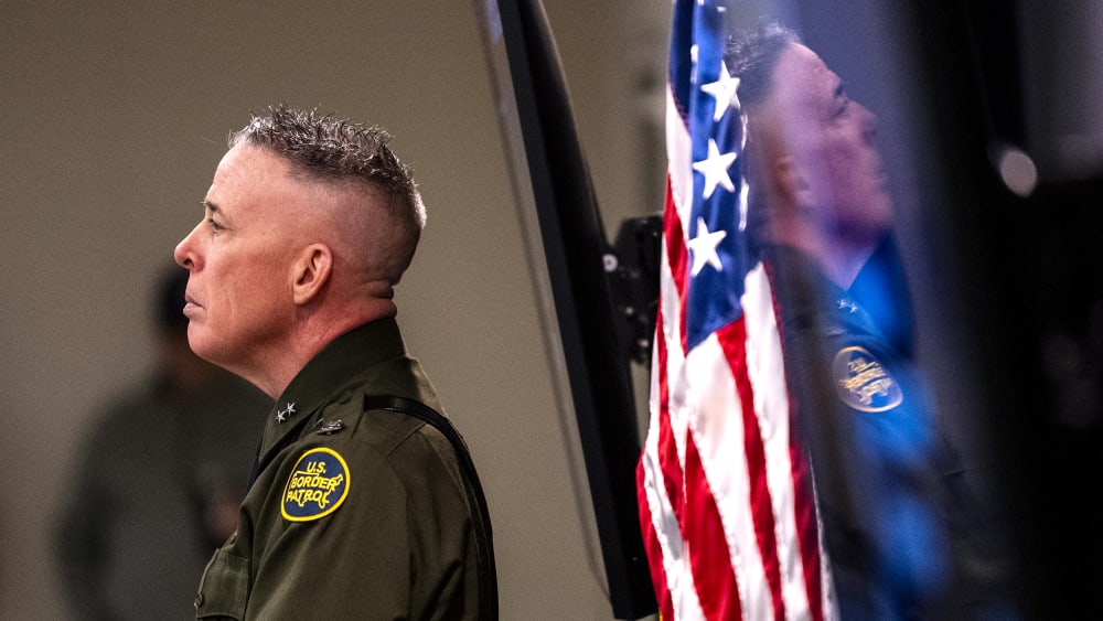 Greg Bovino is seen from the side while he looks on during a presser. His reflection can be seen behind him.