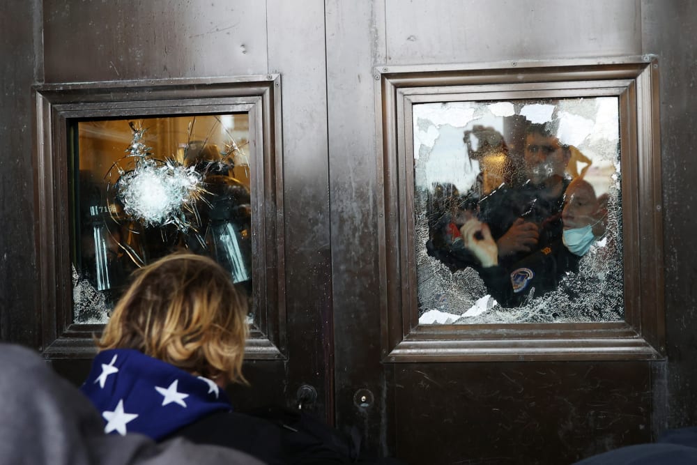 Police officer looks through a broken glass window as protesters stand outside.