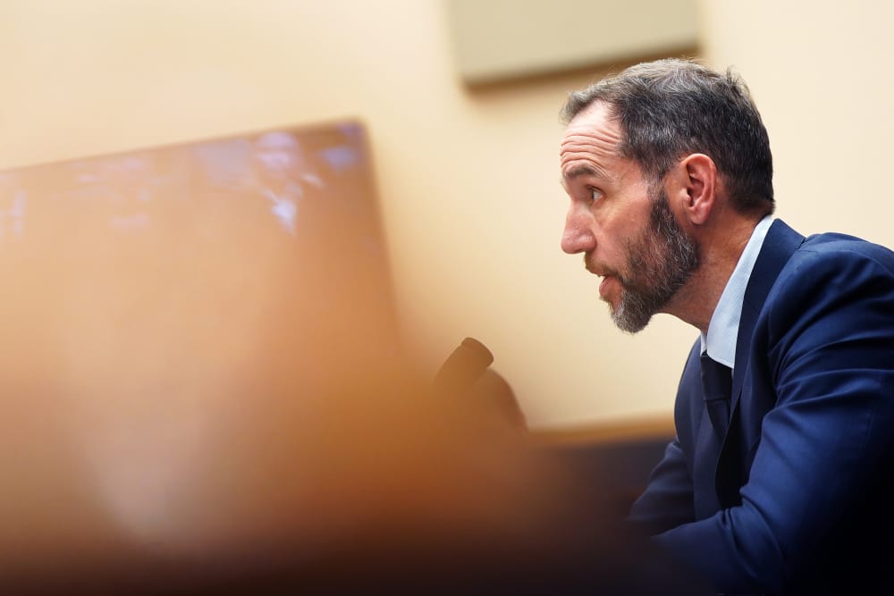 Jack Smith is talking in a tilted frame. What is in front of him is obscured by an out of focus object.