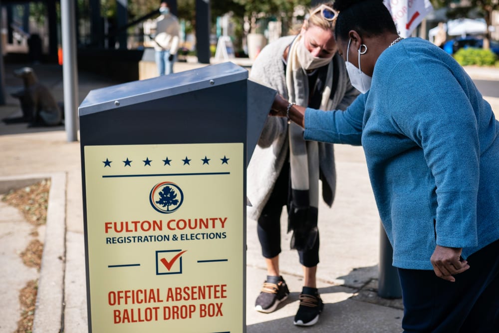 A woman helps another woman to put her absentee ballot into a box outside.
