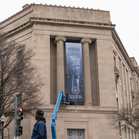 People walk past the Department of Justice building, where construction workers hang a banner of Donald Trump.