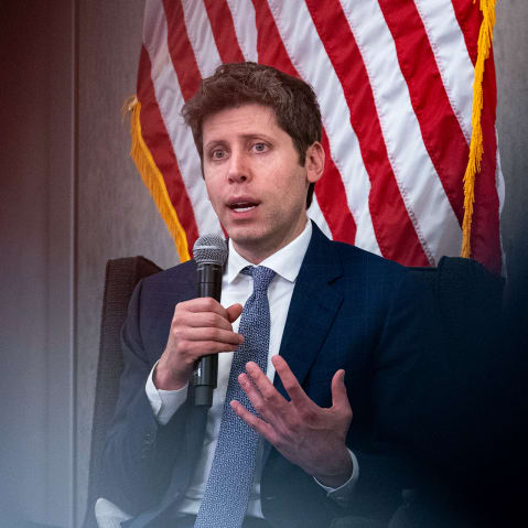 Sam Altman holds a microphone. An American flag is partially visible behind him.
