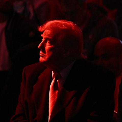 Donald Trump looks into the distance. He and the crowd are basked in red light.