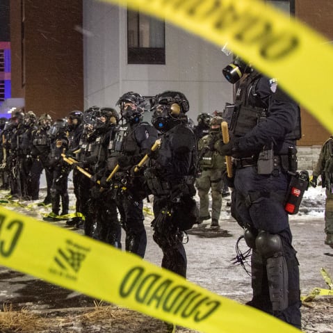 People and law enforcement stand behind strands of yellow caution tape.