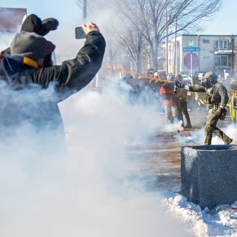 A person on the left films while a federal agent, center, sprays an orange substance ahead. On the right, other agents standby.