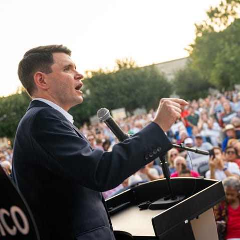 James Talarico speaks at a podium. A crowd of people sits in front of him in a lawn.