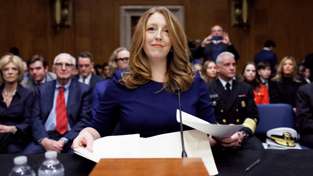 Casey Means sits at a desk, looking ahead and holding papers in her hands.